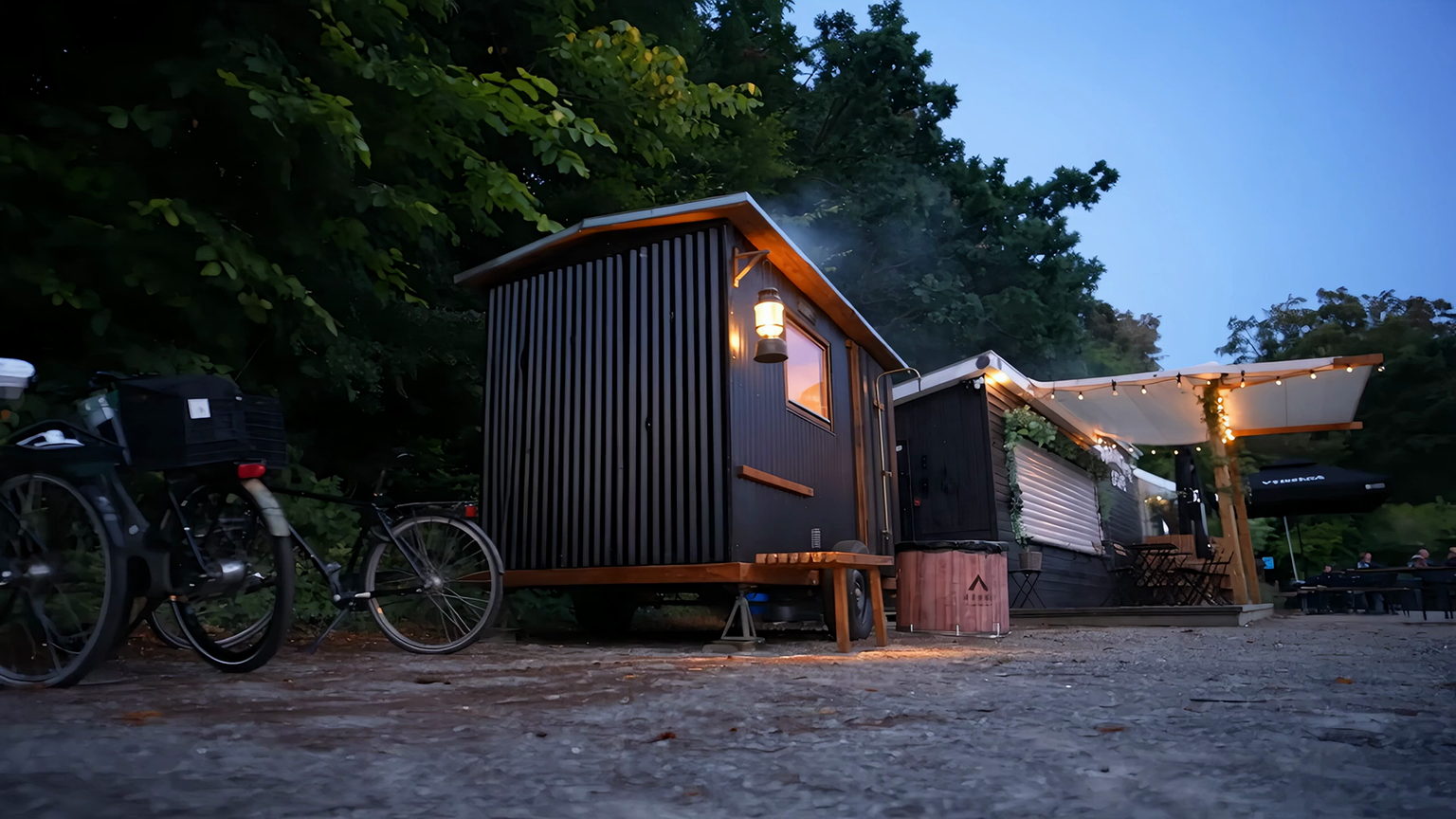 Mobil sauna ved Den Permanente i Risskov — sort bølgeblik-vogn med tændt lanterne og varmt lys i vinduet i blåtimen